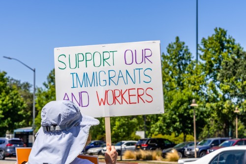 Photo of person wearing a hat protesting with sign that reads: Support our Immigrants and Workers