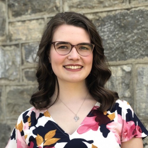 Emily Emick smiling in a floral top in front of a gray brick background.