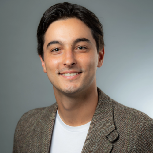 Photo of Adi Jahic wearing a white tee shirt and brown blazer on a plain gray background