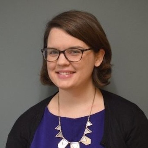 Photo of Phoebe Sloane, who is a caucasian woman with shoulder-length brown hair, wearing brown glasses, a purple shirt, black cardigan, and white chunky necklace