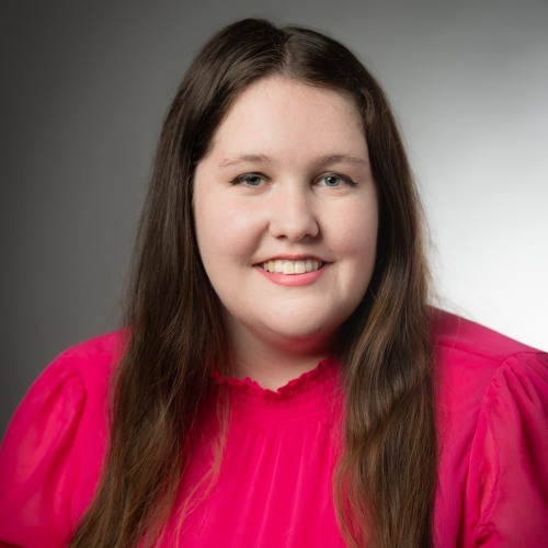 Photo of Rachel Cummings, caucasian woman with long brown hair wearing a bright pink blouse against a gray background