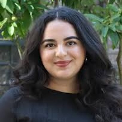 Photo of Diana Gandara who has long black hair and is wearing a black top while standing in front of an outdoor area with trees