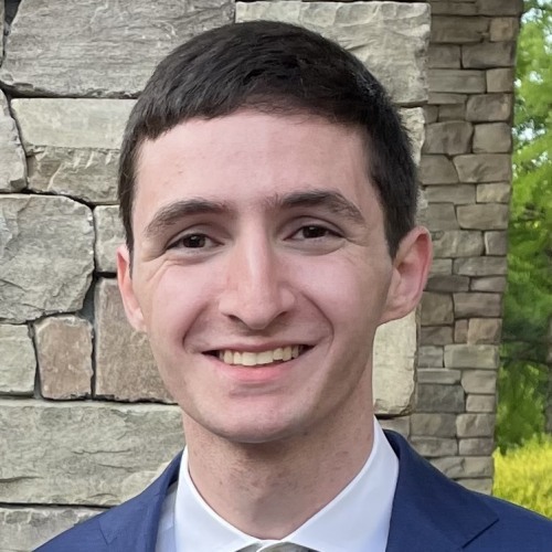 Photo of Maxwell Grozovsky who has short brown hair and is wearing a white button-down shirt under a navy blue blazer and standing against a stone wall background