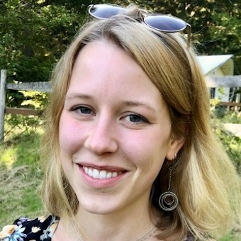 Photo of Mira Guth who has long blonde hair and is wearing a flowered blouse and sunglasses on her head, standing in a park area
