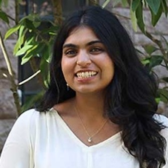 Photo of Bhumi Shah, woman with long brown hair wearing white sweater against a greenery background