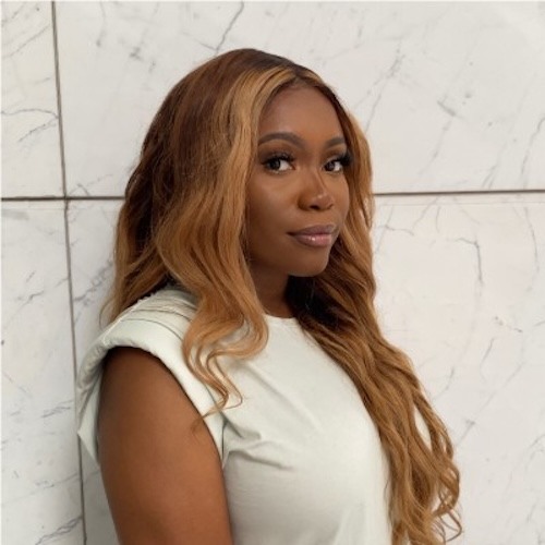 Photo of Kristen Ukeomah who has long brown hair and is wearing a white sleeveless top against a white tiled background