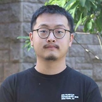 Photo of Yan Zhou who has short brown hair, round framed glasses, and is wearing a black tee shirt while standing against an outdoor background