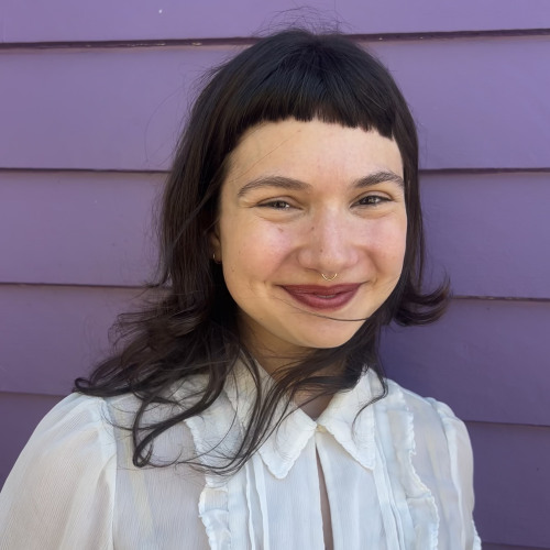 Isabel Levin who has long dark hair with bangs and is wearing a white blouse while standing in front of a purple house