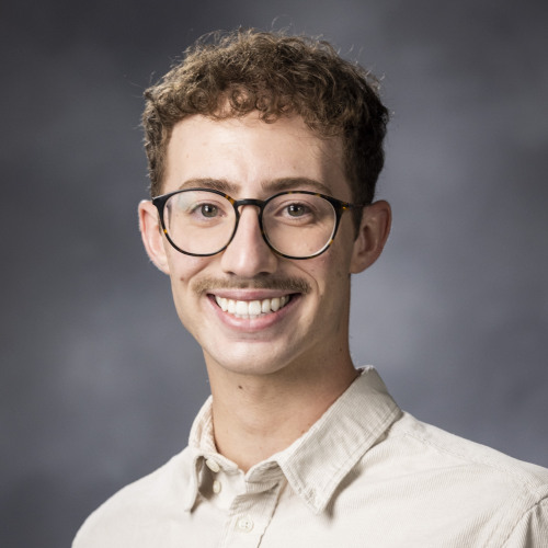 Deegan Minchey who has short brown hair and glasses and is wearing a beige collared shirt and standing in front of a gray background