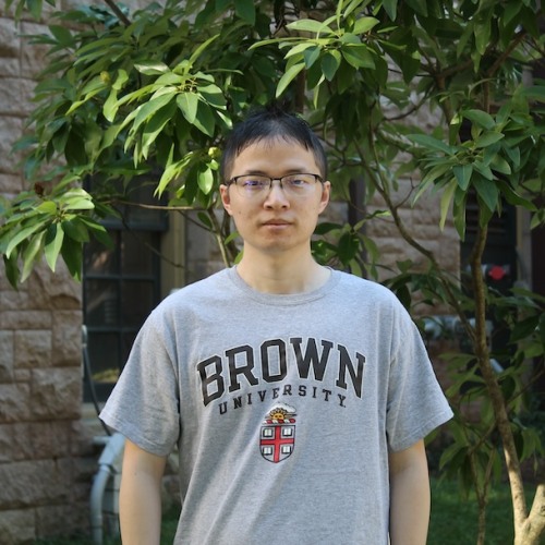 Ang Li wearing Brown t-shirt against brick building and greenery backdrop