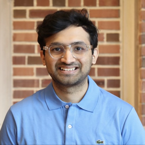 Sharad Hotha wearing blue button down shirt and glasses standing against brick wall