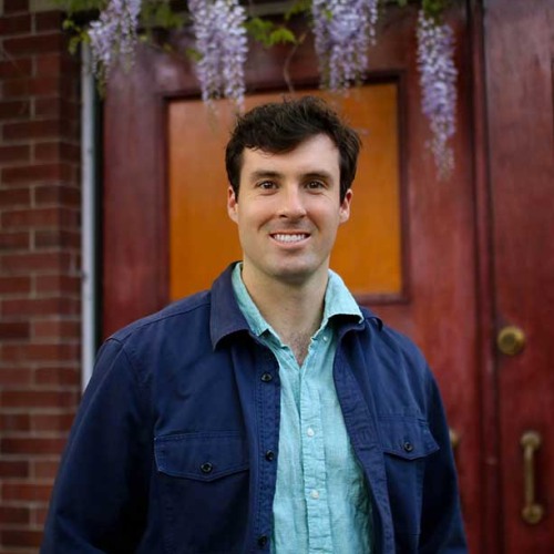 Daniel Krugman wearing blue print shirt and navy blue jacket against brown building backdrop